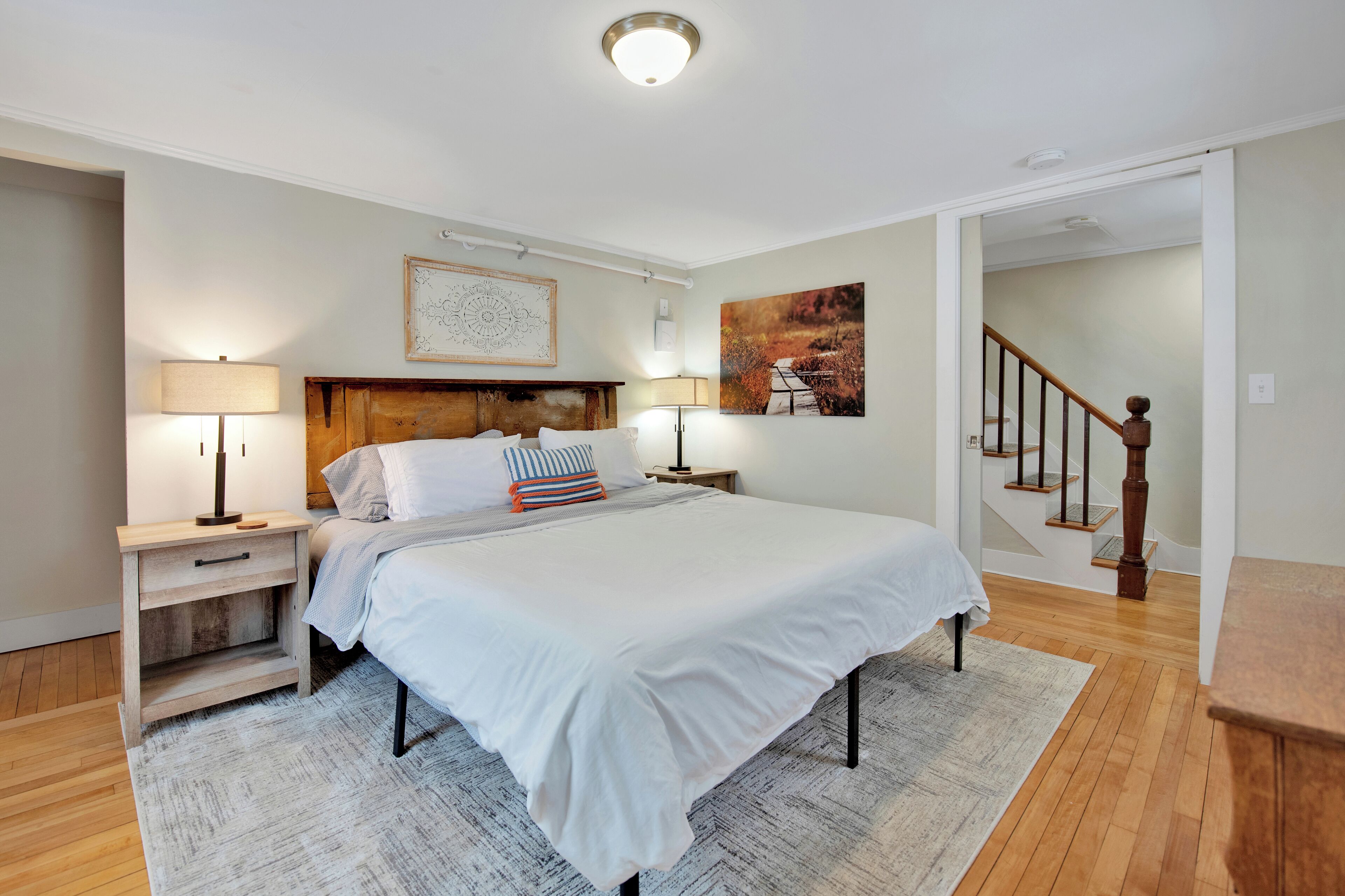 A bedroom with a large bed, two bedside tables with lamps, a wooden headboard, and a staircase in the background.