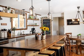 A modern kitchen with wooden countertops, a central island with stools, hanging pendant lights, and a chalkboard menu.