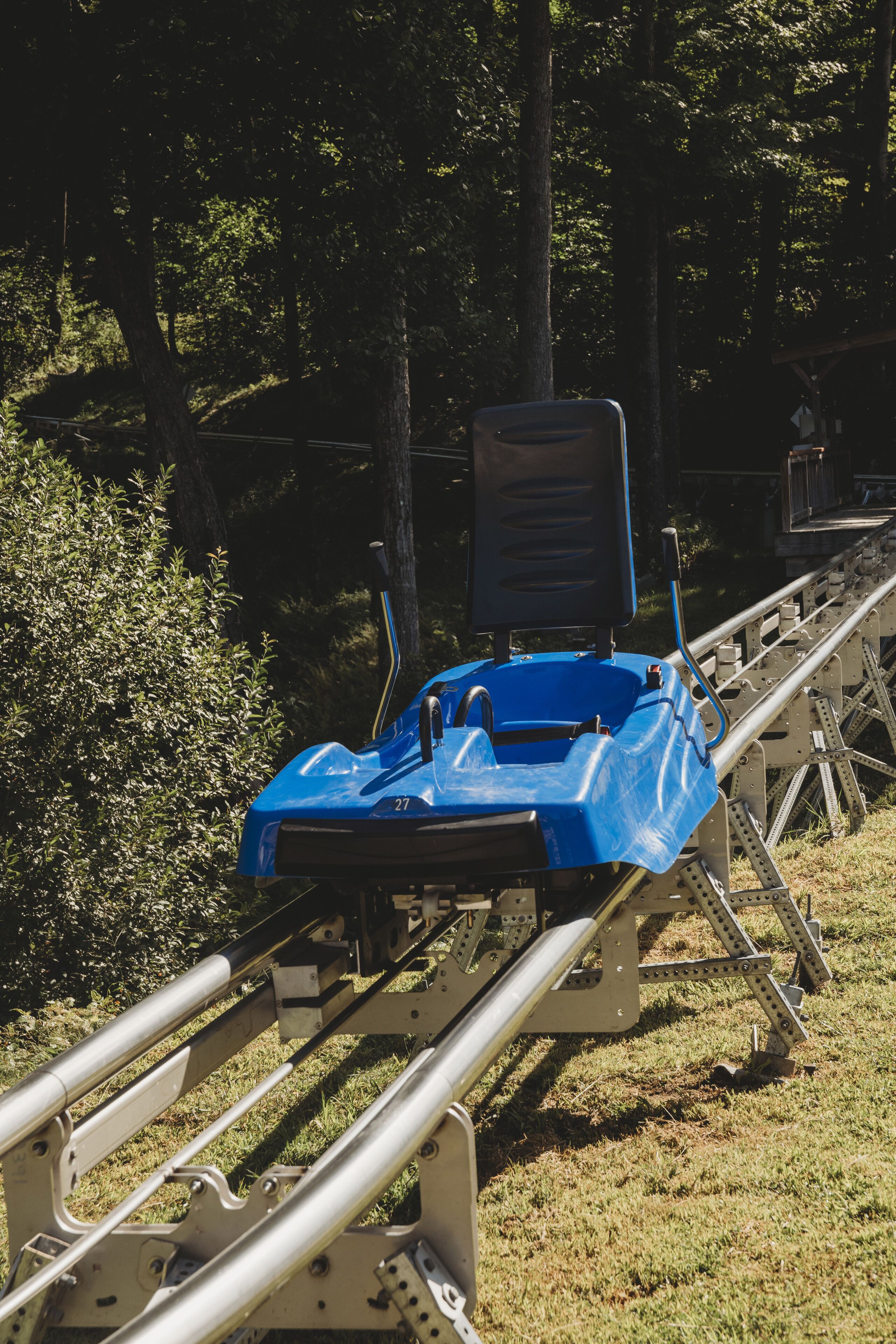 Un vehículo azul sobre una vía con soportes metálicos, rodeado de árboles y césped.