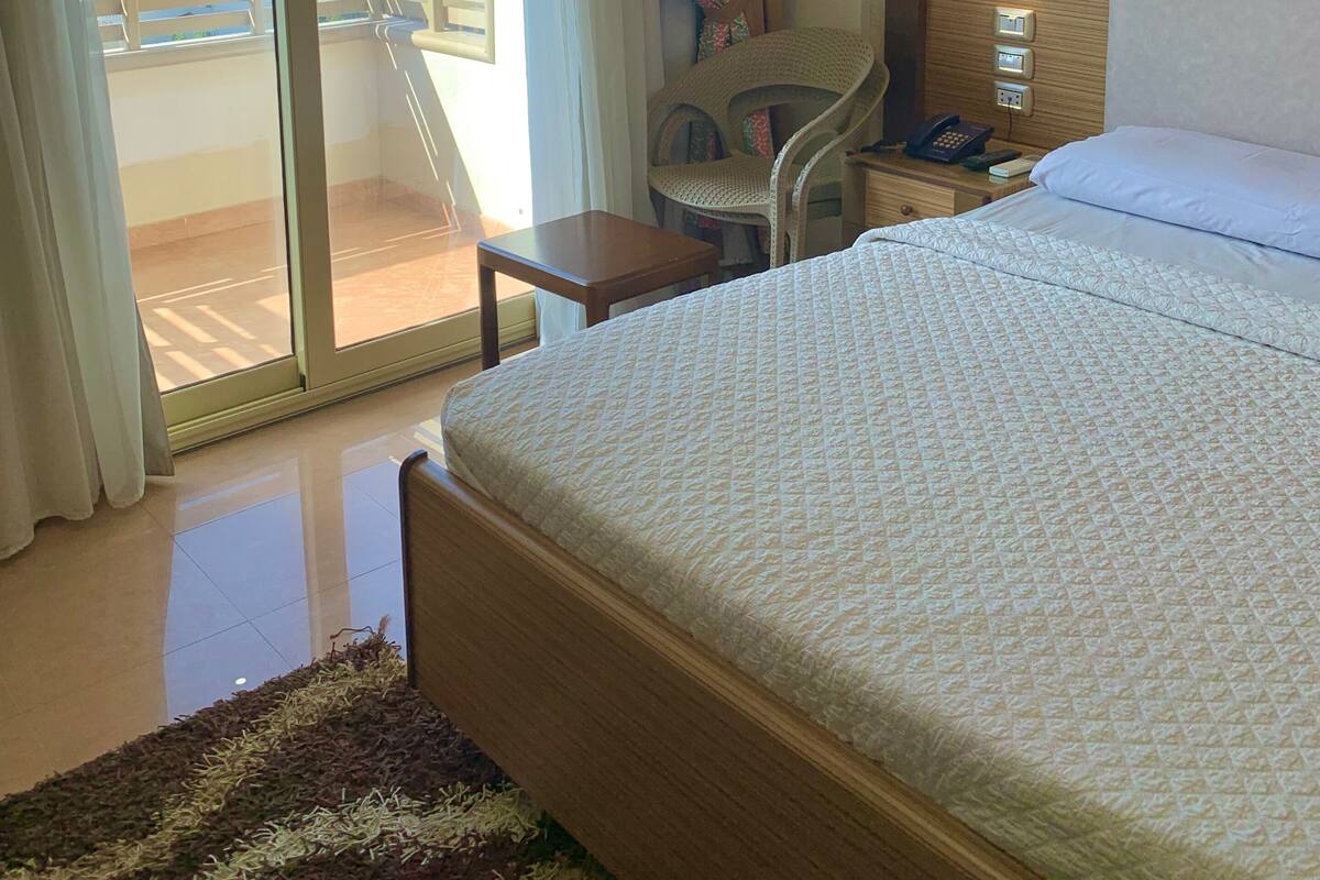 Habitación de hotel con cama, una silla, una mesita, una mesita de noche con teléfono y vistas al exterior.