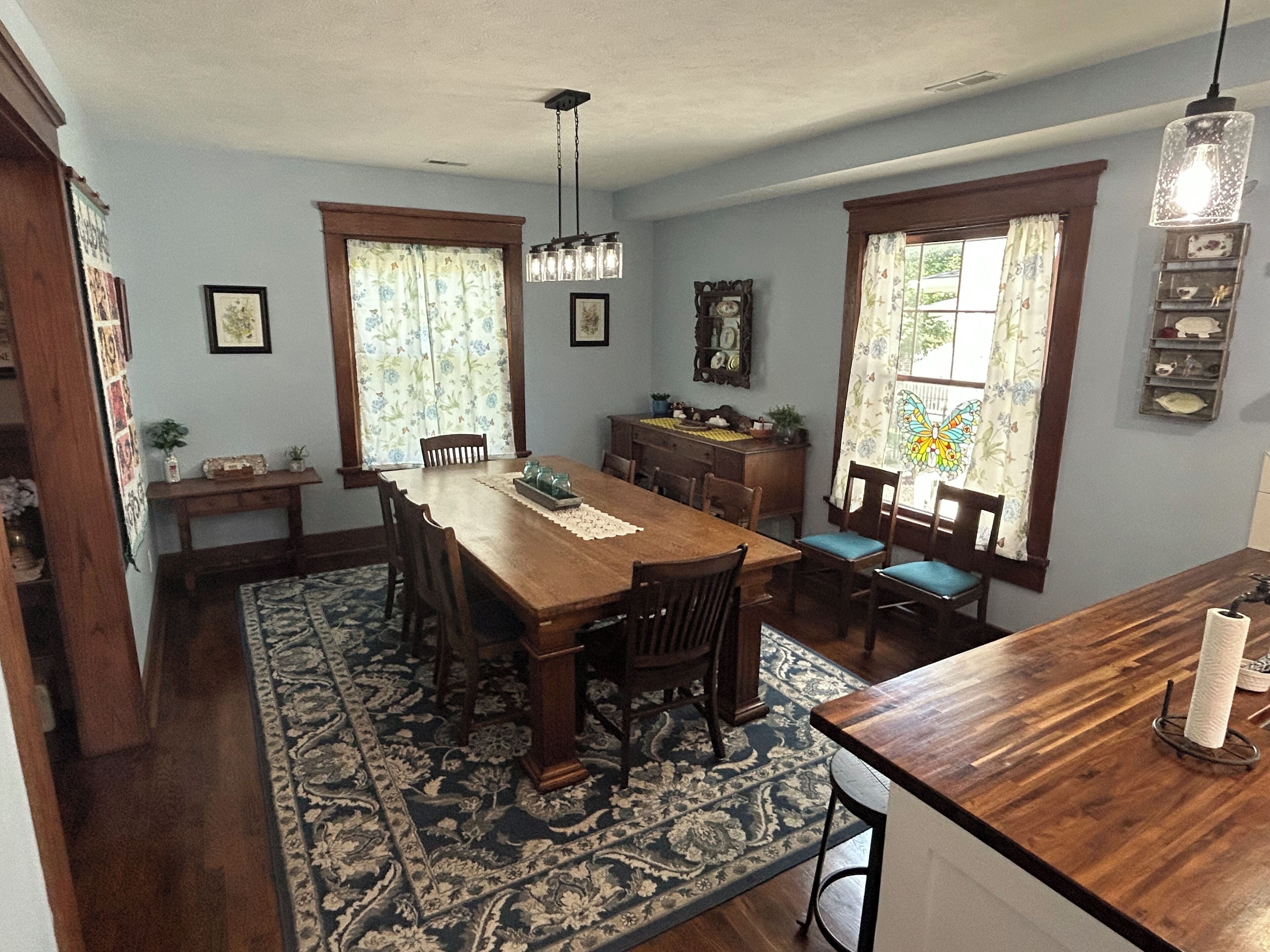 Une salle à manger avec une table en bois, des chaises, un lustre et un tapis à motifs.