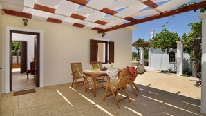 Une terrasse couverte avec une table et des chaises en bois, un sol carrelé et un mur blanc agrémenté d’une fenêtre et de volets.