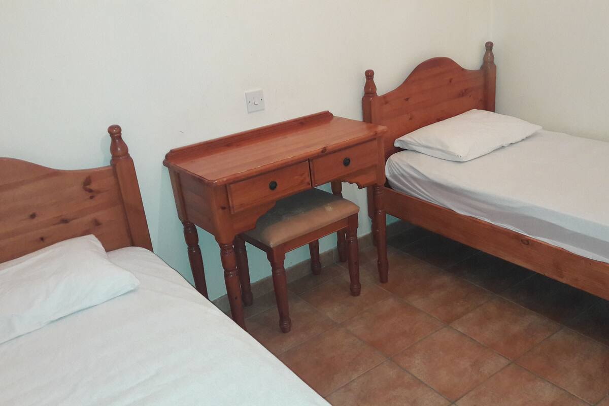 A bedroom with two wooden beds, a wooden nightstand with a drawer, and a small bench.