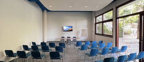 Una sala conferenze con file di sedie blu, un grande schermo e vista sull'esterno grazie a ampie finestre.
