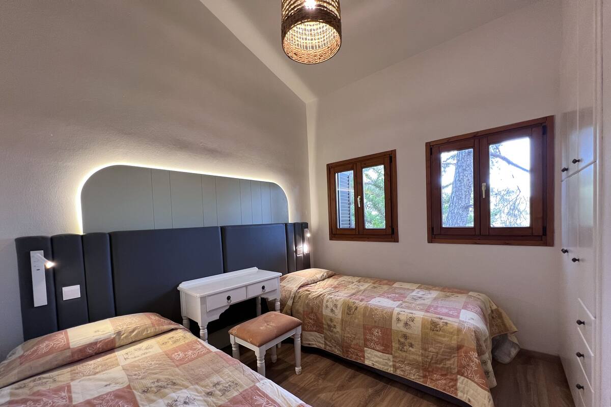 A bedroom with two beds, a bedside table, a small bench, and two windows.