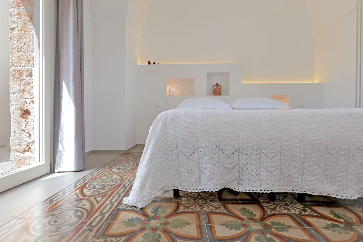 A bedroom with a bed, a patterned floor, a window with curtains, and wall-mounted lights.