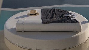 Vista desde una ventana de avión de un barco en el agua, una cama redonda blanca con un sombrero y un libro, y un paisaje montañoso al fondo.
