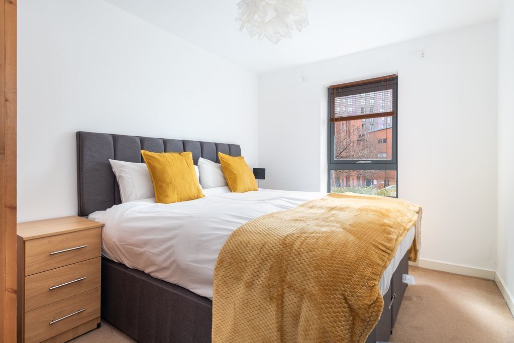 Une chambre à coucher comprenant un lit, une table de chevet, une fenêtre donnant sur des immeubles et une armoire en bois.