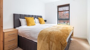 A bedroom with a bed, a nightstand, a window with a view of buildings, and a wooden wardrobe. - Bouverie Apartment (Leeds)