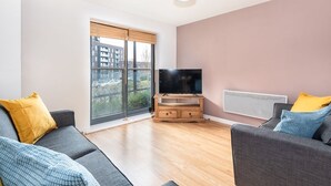 A modern living room with a large window, a flat-screen TV on a wooden stand, and two grey sofas with yellow and blue cushions. - Bouverie Apartment (Leeds)