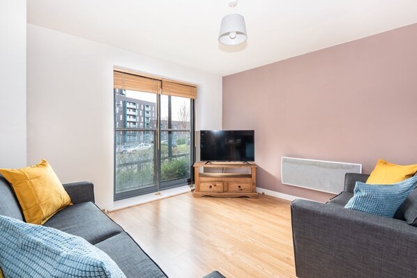 A modern living room with a large window, a flat-screen TV on a wooden stand, and two grey sofas with yellow and blue cushions. - Bouverie Apartment (Leeds)