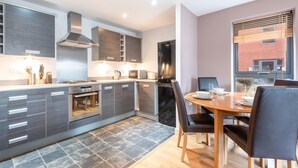 A modern kitchen with grey cabinetry, a built-in oven, and a dining area with a wooden table and chairs. - Bouverie Apartment (Leeds)