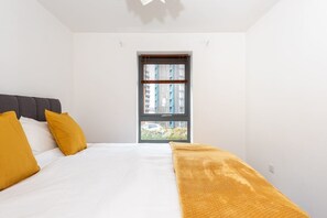 A bedroom with a bed, two pillows, a blanket, and a window with blinds. - Bouverie Apartment (Leeds)