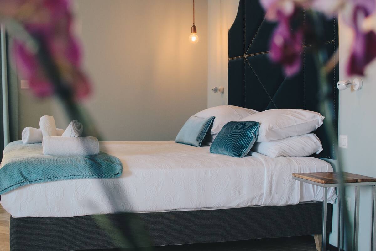 Una cama bien tendida con sábanas blancas y un plaid verde azulado, una mesita de noche con una lámpara y un cabecero oscuro.