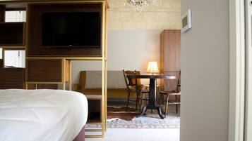 A hotel room with a bed, a television mounted on a wooden panel, a dining table with chairs, and a chandelier.