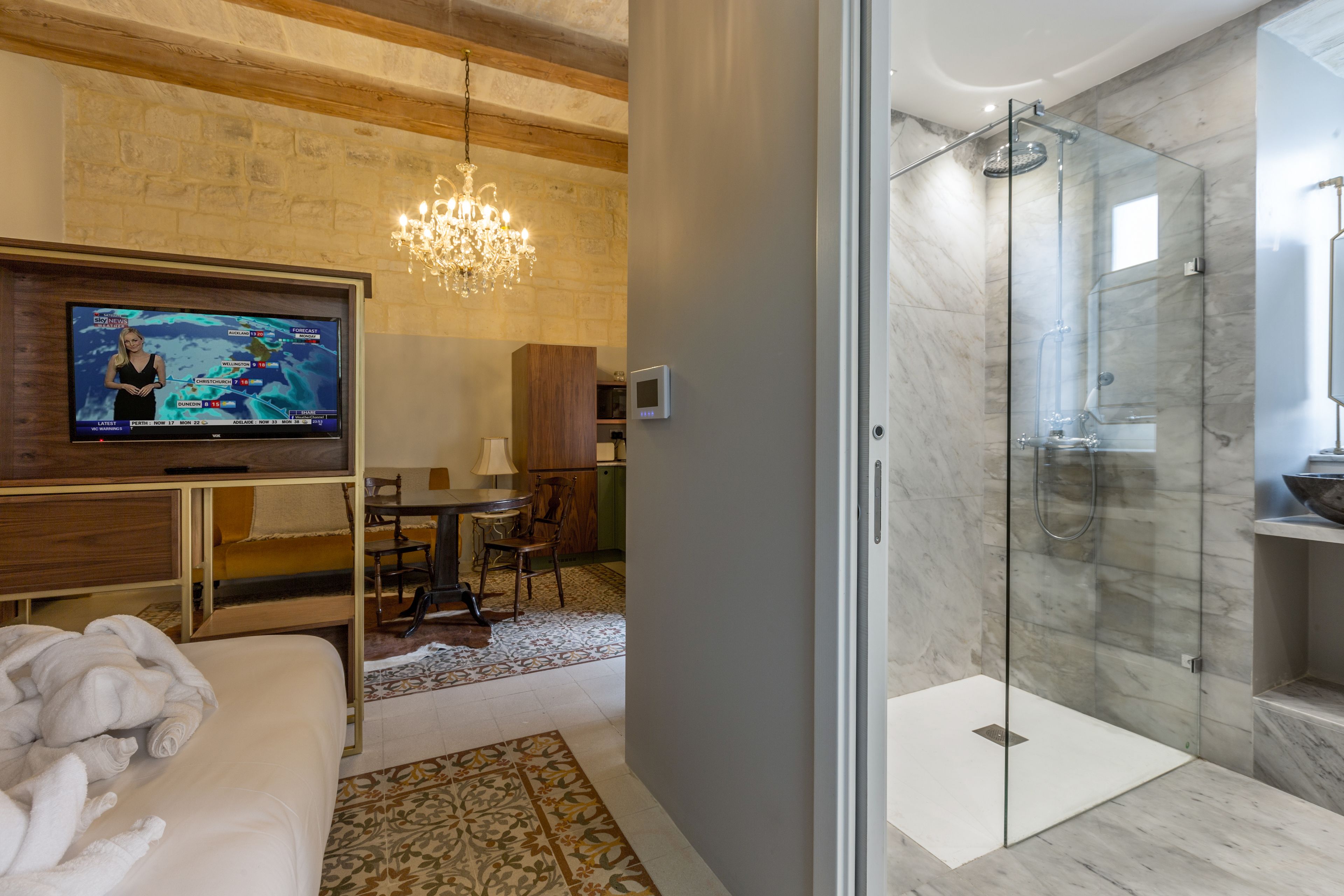 Ein modernes Hotelzimmer mit Fernseher, einer in Glas abgeschirmten Dusche, Holzboden und einem Kronleuchter.