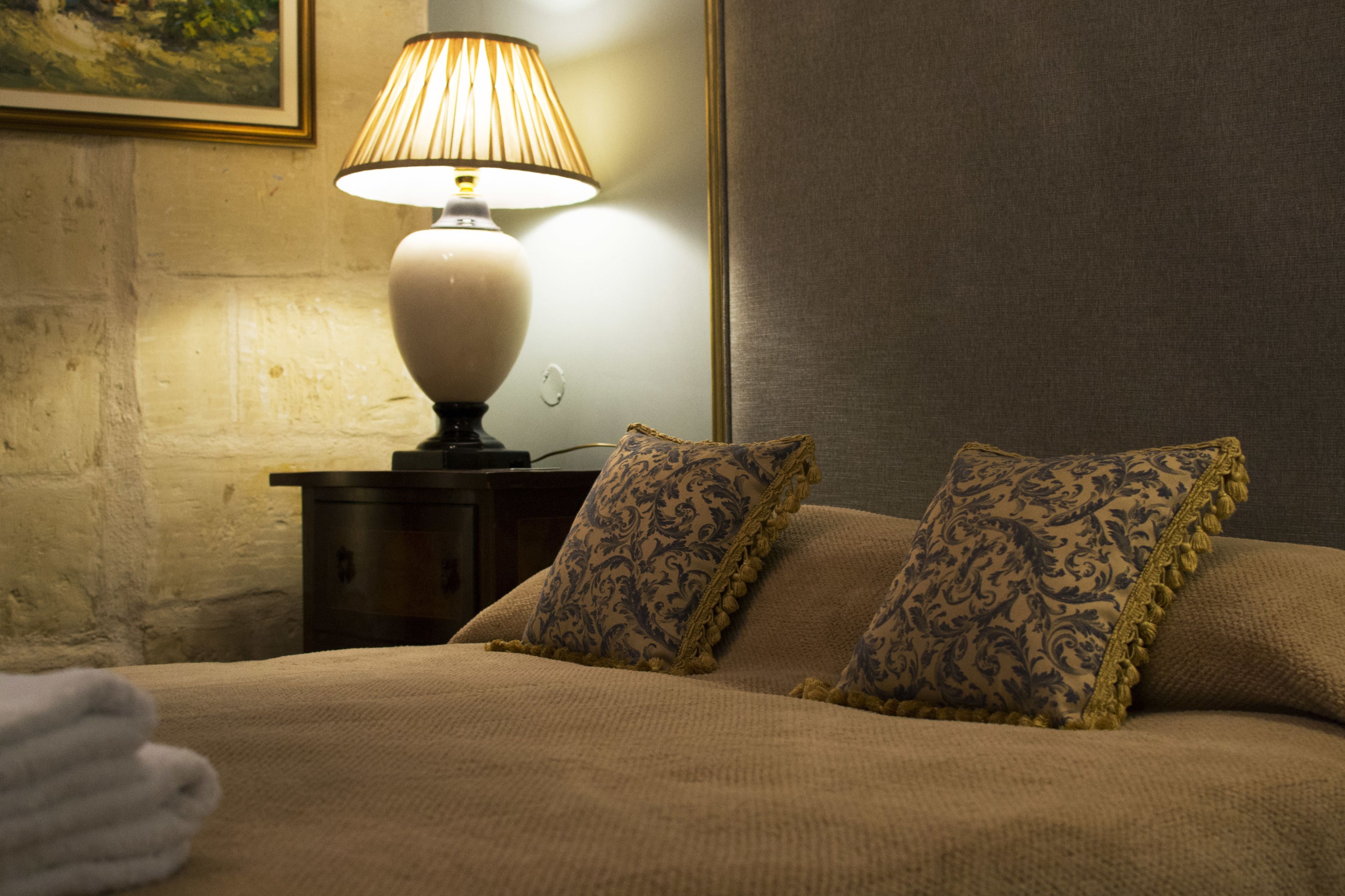 Un lit bien fait, recouvert d’une couverture beige et agrémenté de deux coussins à motifs, une lampe de chevet et un tableau encadré au mur.
