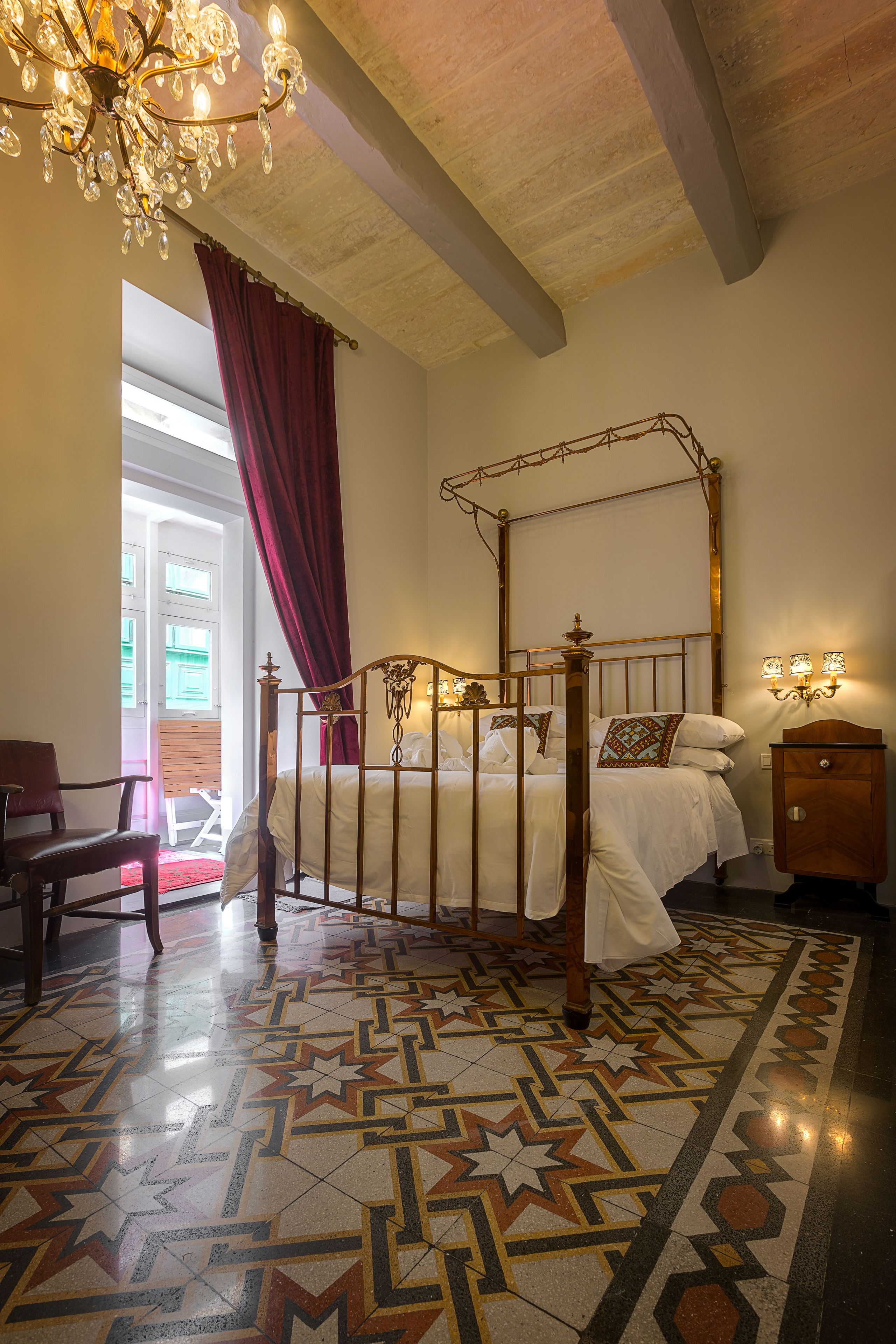 A bedroom with a four-poster bed, a chandelier, a patterned floor, and a wooden nightstand.