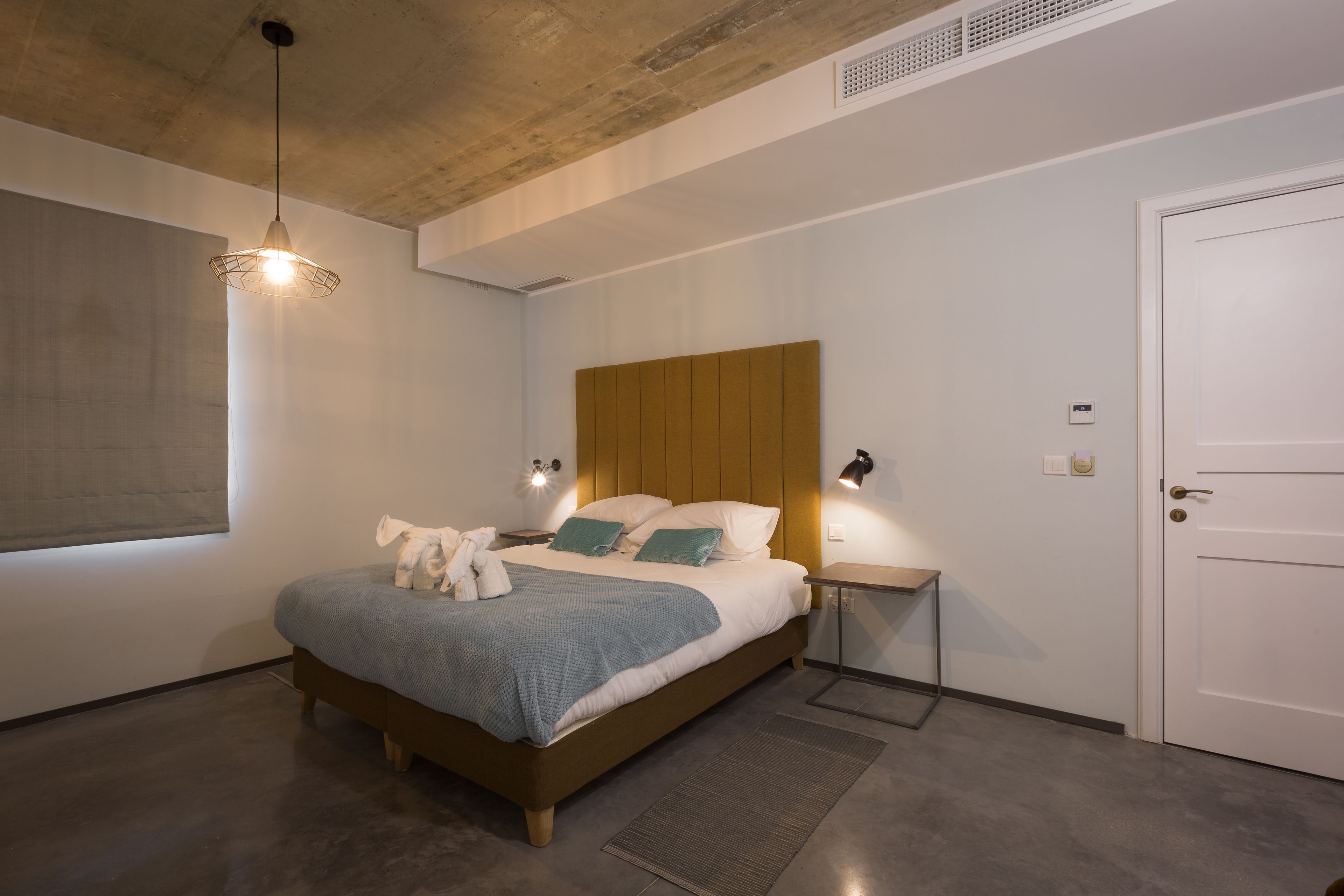Une chambre à coucher moderne avec une tête de lit en bois, un grand lit recouvert d’une couverture bleue, des lampes de chevet et une porte.