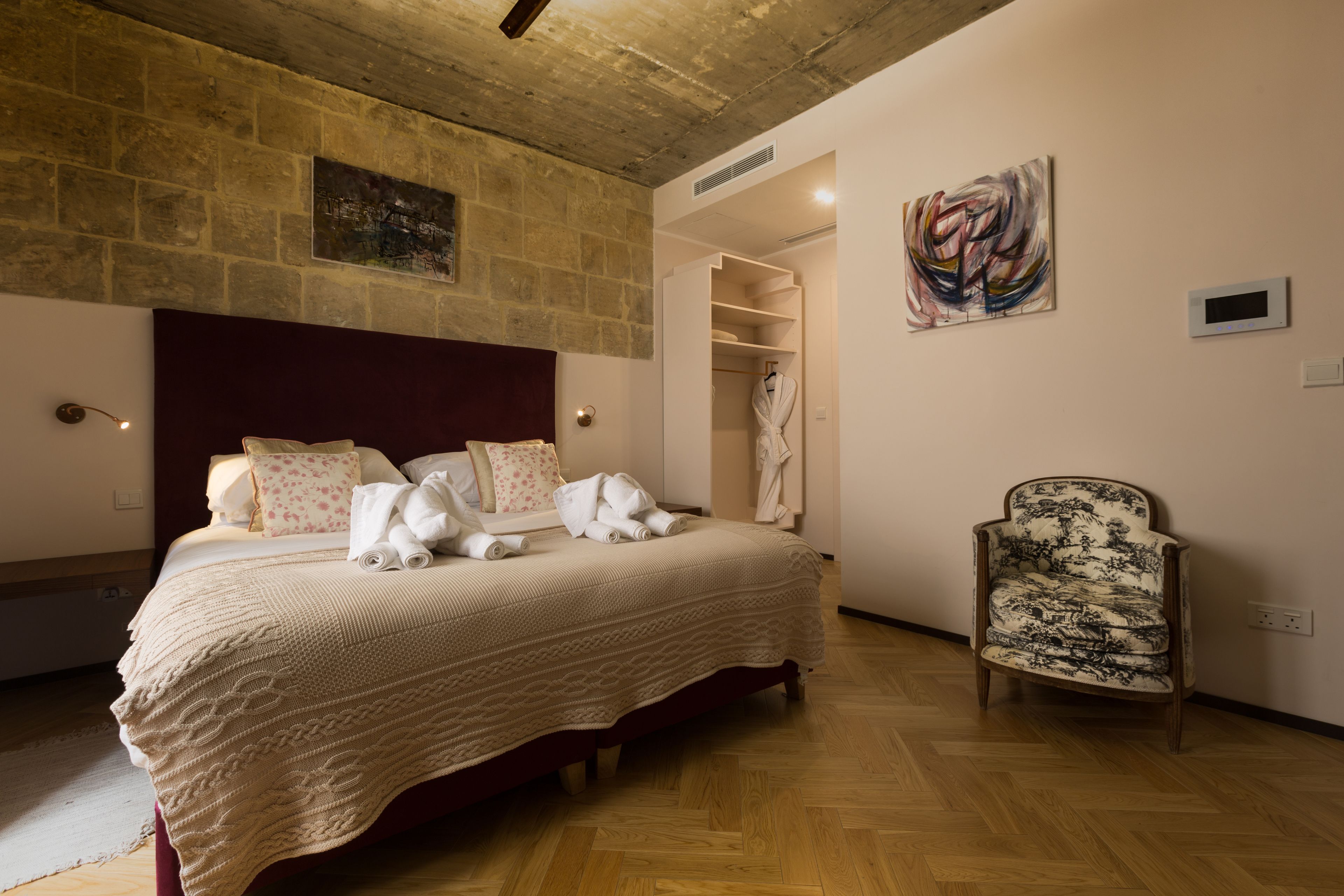 A bedroom with a wooden floor, a large bed with a white bedspread, a wooden headboard, a chair with a patterned cushion, and a painting on the wall.
