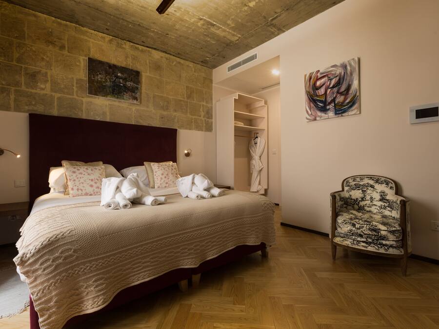 Una camera da letto con pavimento in legno, un letto grande con copripiumino bianco, una testiera in legno, una sedia con cuscino decorato e un quadro appeso al muro.