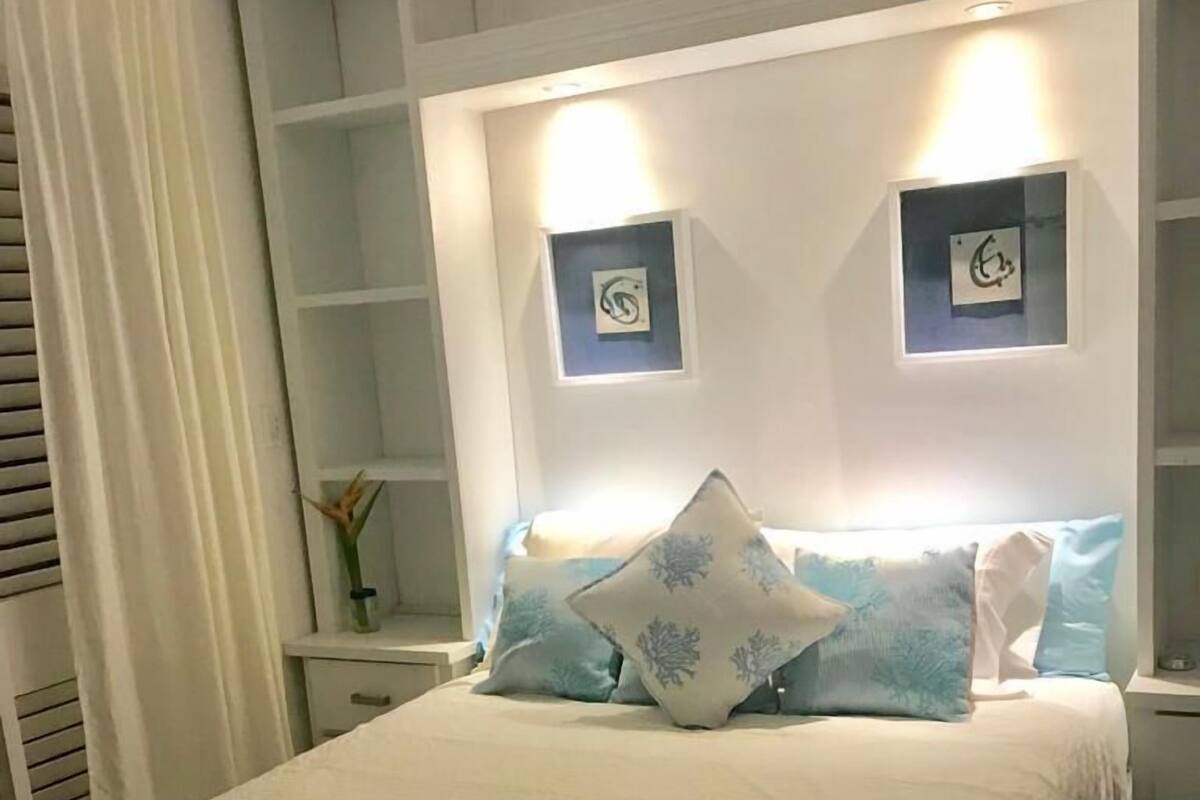 A neatly made bed with a blue-striped blanket, a white headboard with two wall-mounted lights, and a small bedside table with a vase of flowers.