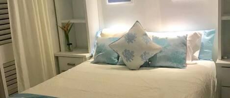 A neatly made bed with a blue-striped blanket, a white headboard with two wall-mounted lights, and a small bedside table with a vase of flowers.