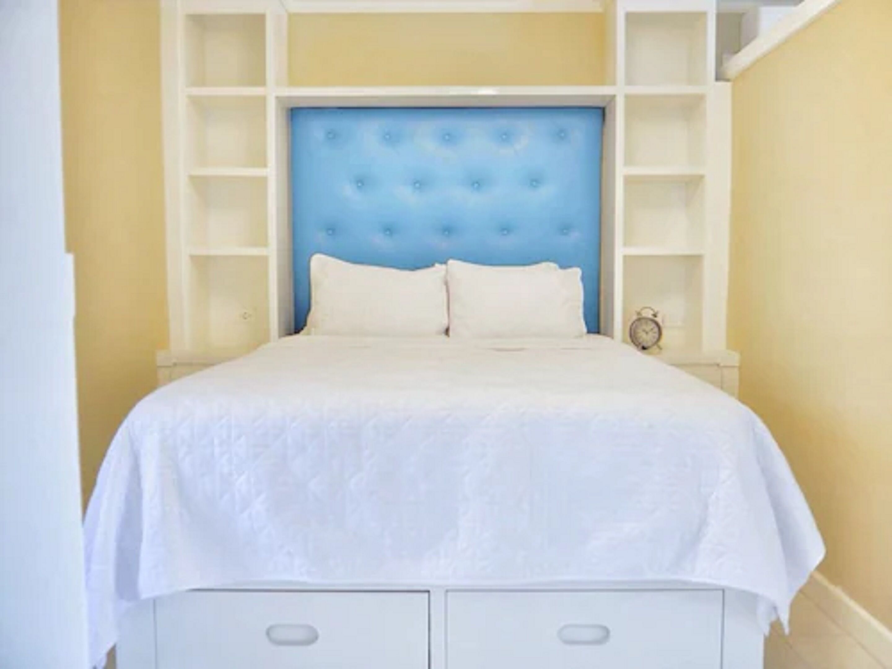 Un dormitorio con un cabecero azul capitonado, ropa de cama blanca y estanterías empotradas.