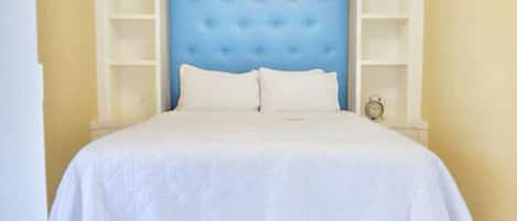 Un dormitorio con un cabecero azul capitonado, ropa de cama blanca y estanterías empotradas.