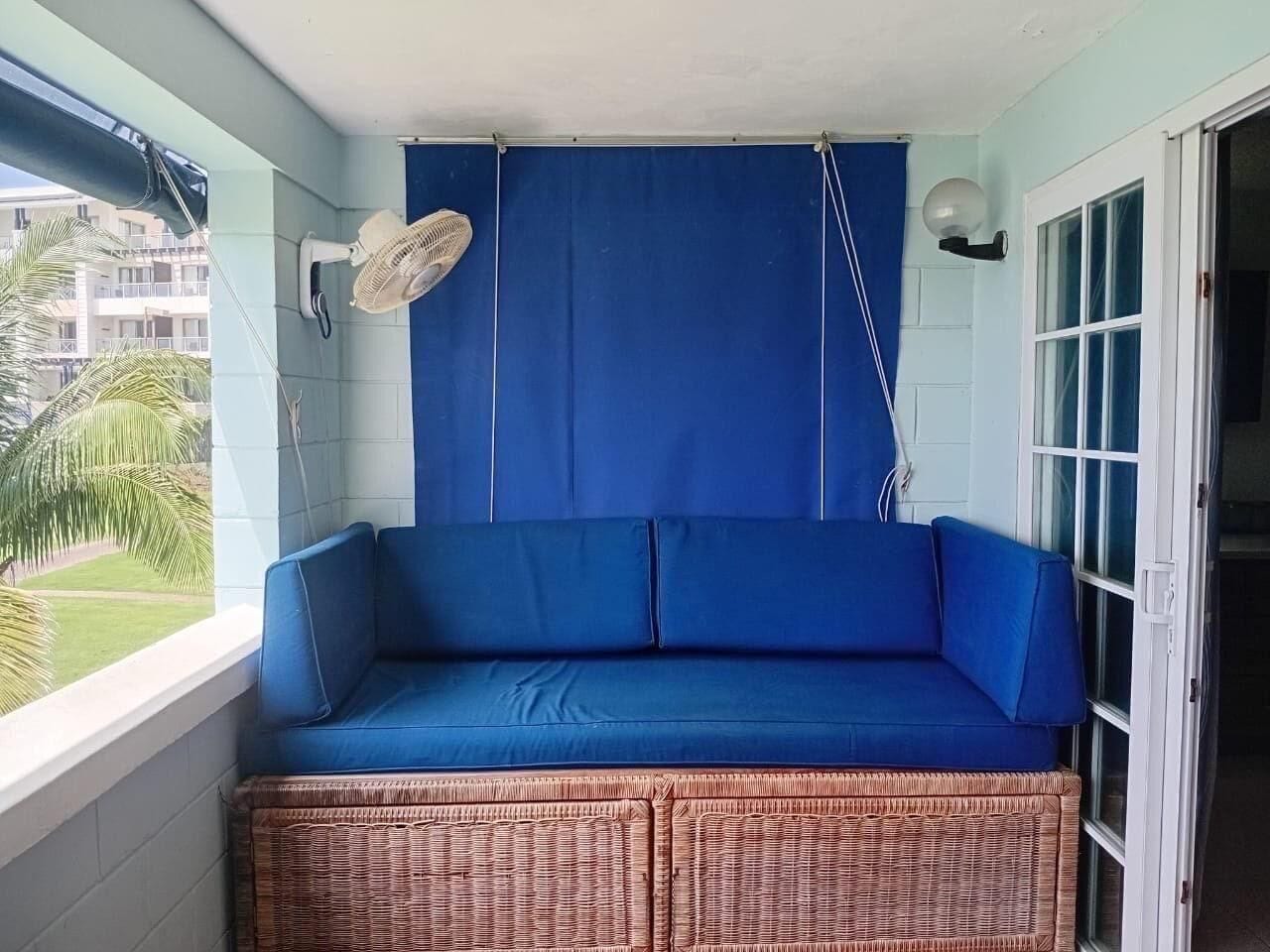 A balcony with a blue cushioned seating area, a fan, and a view of a building and palm trees.
