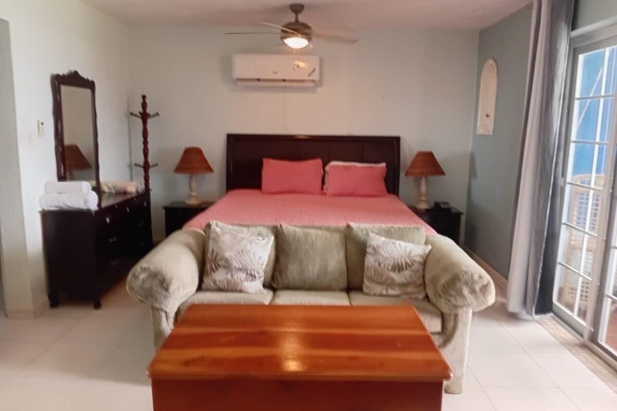 A bedroom with a bed, two bedside tables, a ceiling fan, an air conditioning unit, a window with curtains, and a wooden bench.