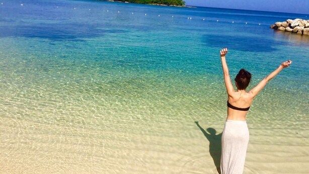 Une personne se tient sur une plage, les bras levés, face à une mer calme et à une petite île qui se dessine à l’horizon.