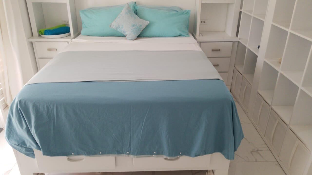 Un lit avec une couvre-lit bleue et des oreillers, une table de chevet blanche sur laquelle se trouve un objet bleu, et une armoire blanche dont les portes sont ouvertes.