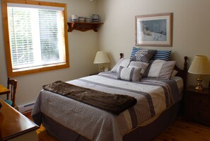 Ein ordentlich bezogenes Bett mit gestreifter Bettdecke und Kissen, ein Nachttisch aus Holz mit einer Lampe, ein Fenster mit Jalousien und ein Regal mit Dekoration.