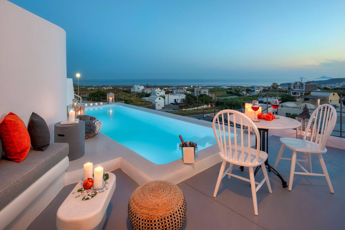 A rooftop terrace with a swimming pool, white chairs, candles, and a view of the sea and buildings.
