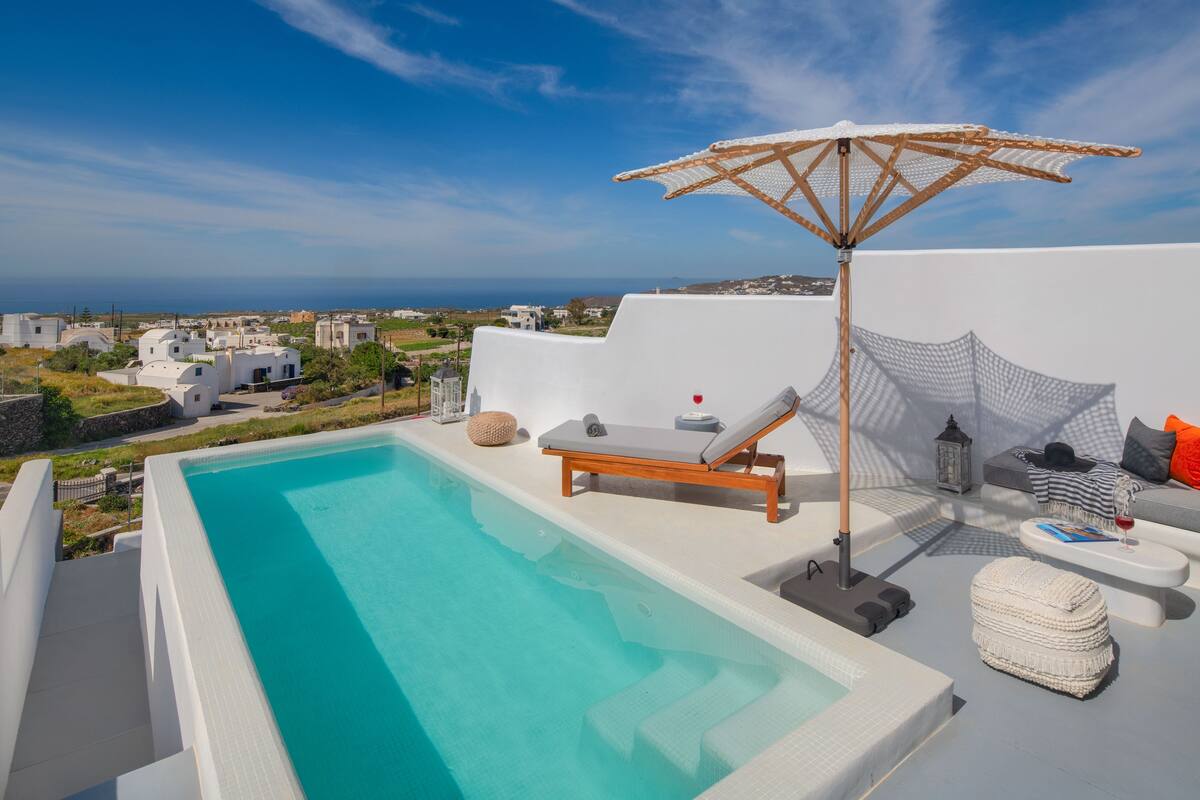 A rooftop pool area with a view of the sea, a lounge chair, a small table with a drink, and a decorative umbrella.