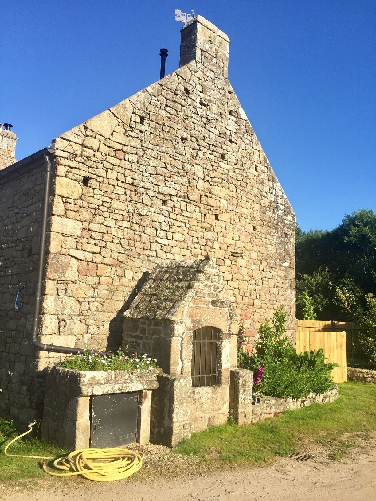 A stone building with a sloped roof, a small window, and a garden area with flowers and plants.