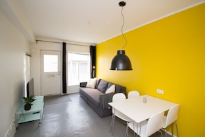 A modern living space with a sofa, a dining table with chairs, a potted plant, and a large window.