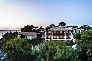 A multi-story hotel with balconies, surrounded by lush greenery and a view of the sea.