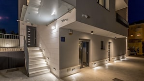 Ein modernes Gebäude mit einer Treppe, die zu einem Eingang führt, einem überdachten Außenbereich und einem bereitstehenden Auto in der Nähe.