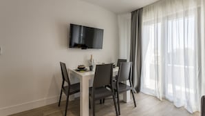 Ein zeitgemäßer Essbereich mit einem weißen Tisch, vier Stühlen, einem an der Wand befestigten Flachbildfernseher und einem großen Fenster mit durchsichtigen Vorhängen.