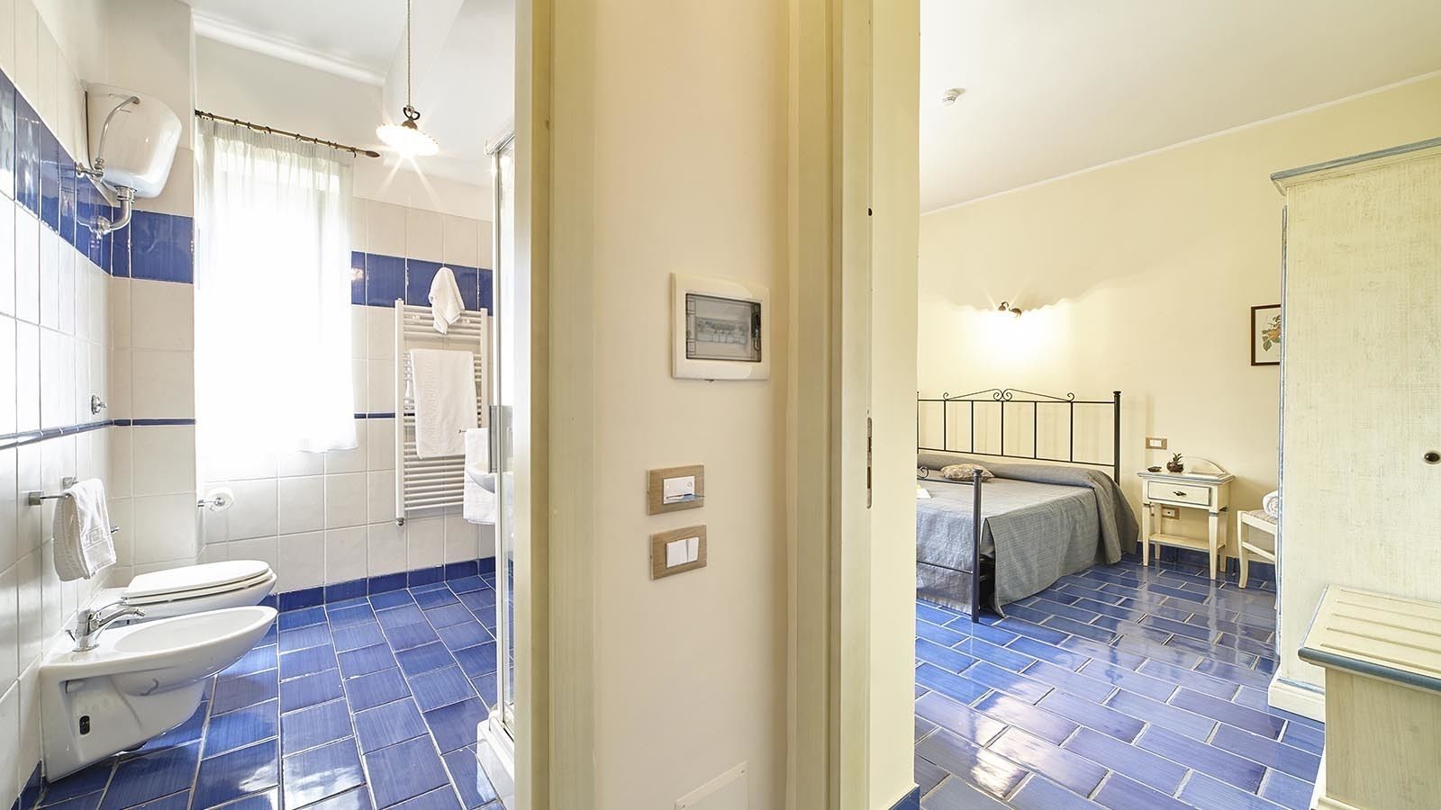 Ein Badezimmer mit Toilette, Waschbecken und Duschbereich. Der Raum hat blaue Fliesenböden und -wände, und im angrenzenden Schlafzimmer befindet sich ein Bett mit Kopfteil.