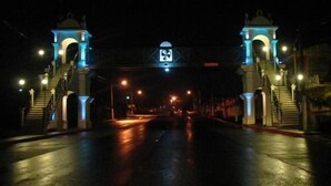 A nighttime view of a street with two illuminated towers at the entrance, a wet road, and a traffic sign overhead.