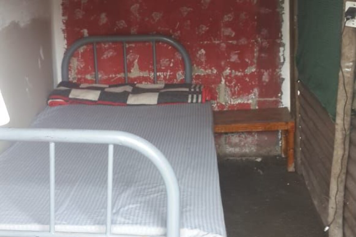 A small, narrow room with a single metal bed, a red wall with peeling paint, and a green door.