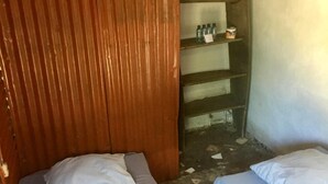 A small, rustic room with two single beds, wooden paneling on one wall, and a shelf with bottles and a container.