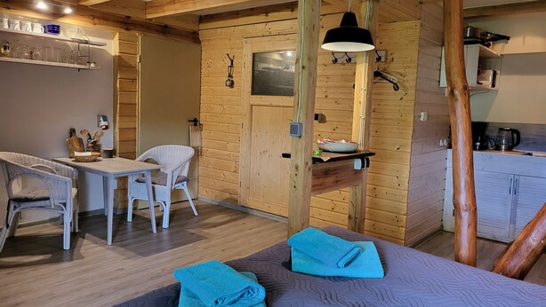 Um quarto acolhedor com paredes de madeira, um espaço de jantar com mesa e cadeiras, e uma cama com toalhas dobradas.