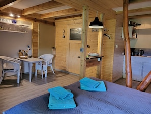 Une pièce chaleureuse aux murs en bois, un coin repas avec une table et des chaises, et un lit avec des serviettes pliées.