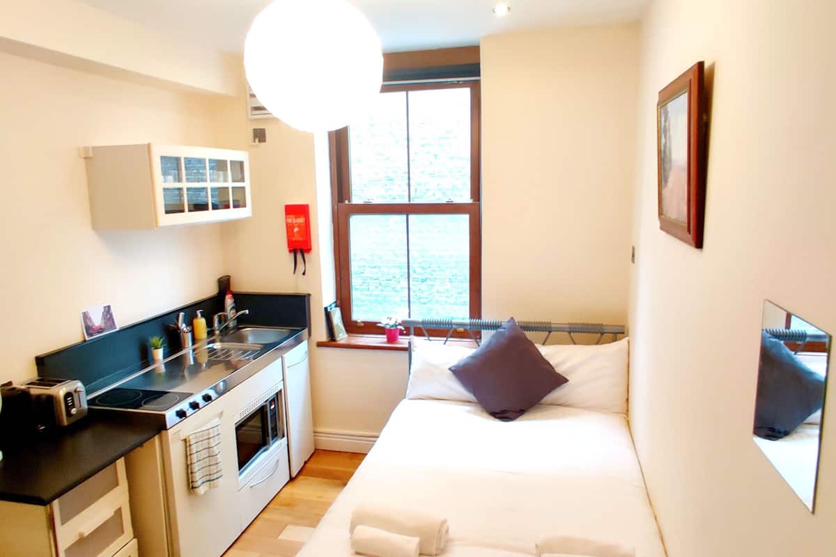 A compact hotel room with a kitchenette, a bed, a window, and a mirror.