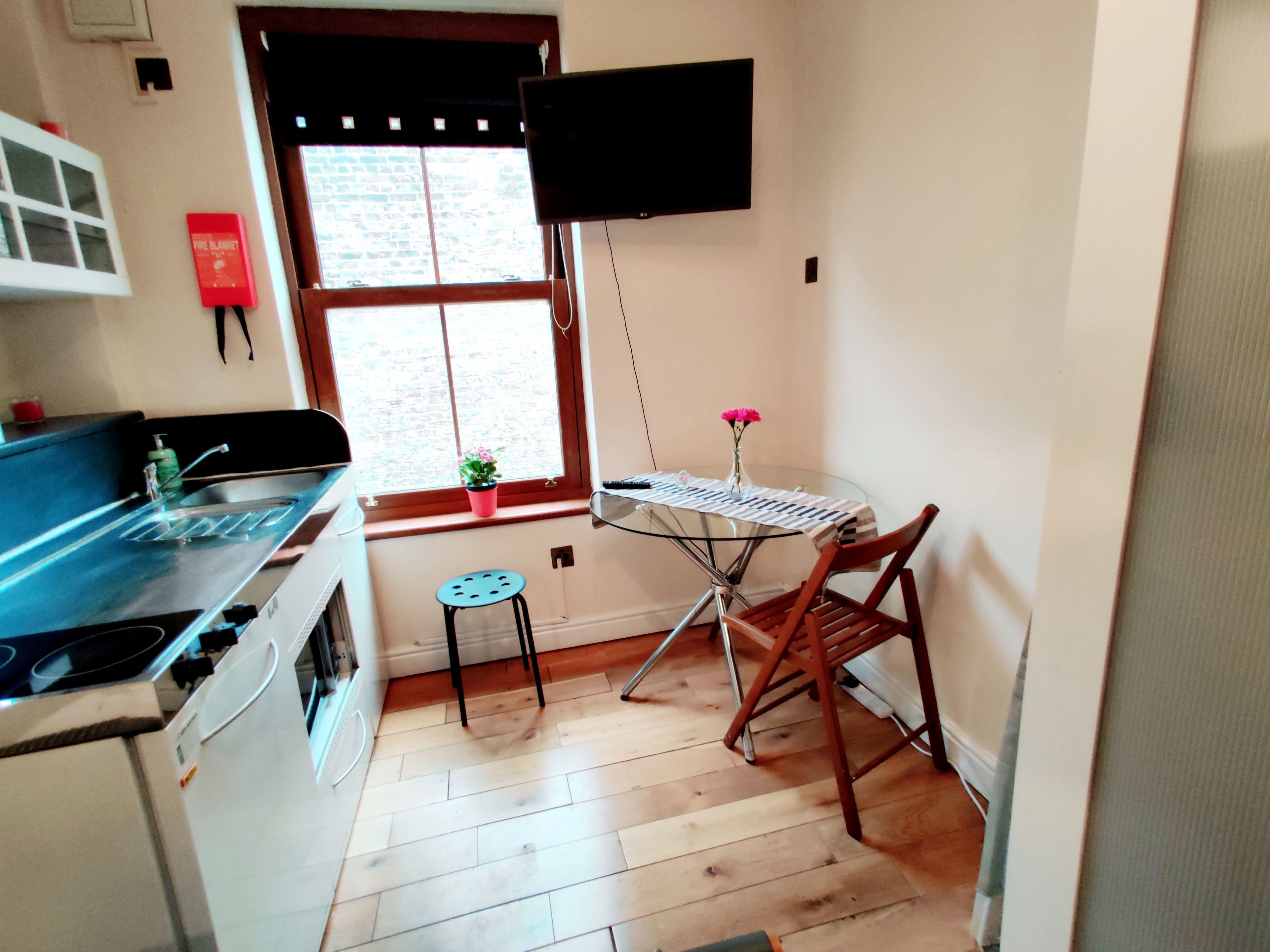 A kitchen with a stove, a sink, a small stool, a table with a keyboard, and a TV mounted on the wall.