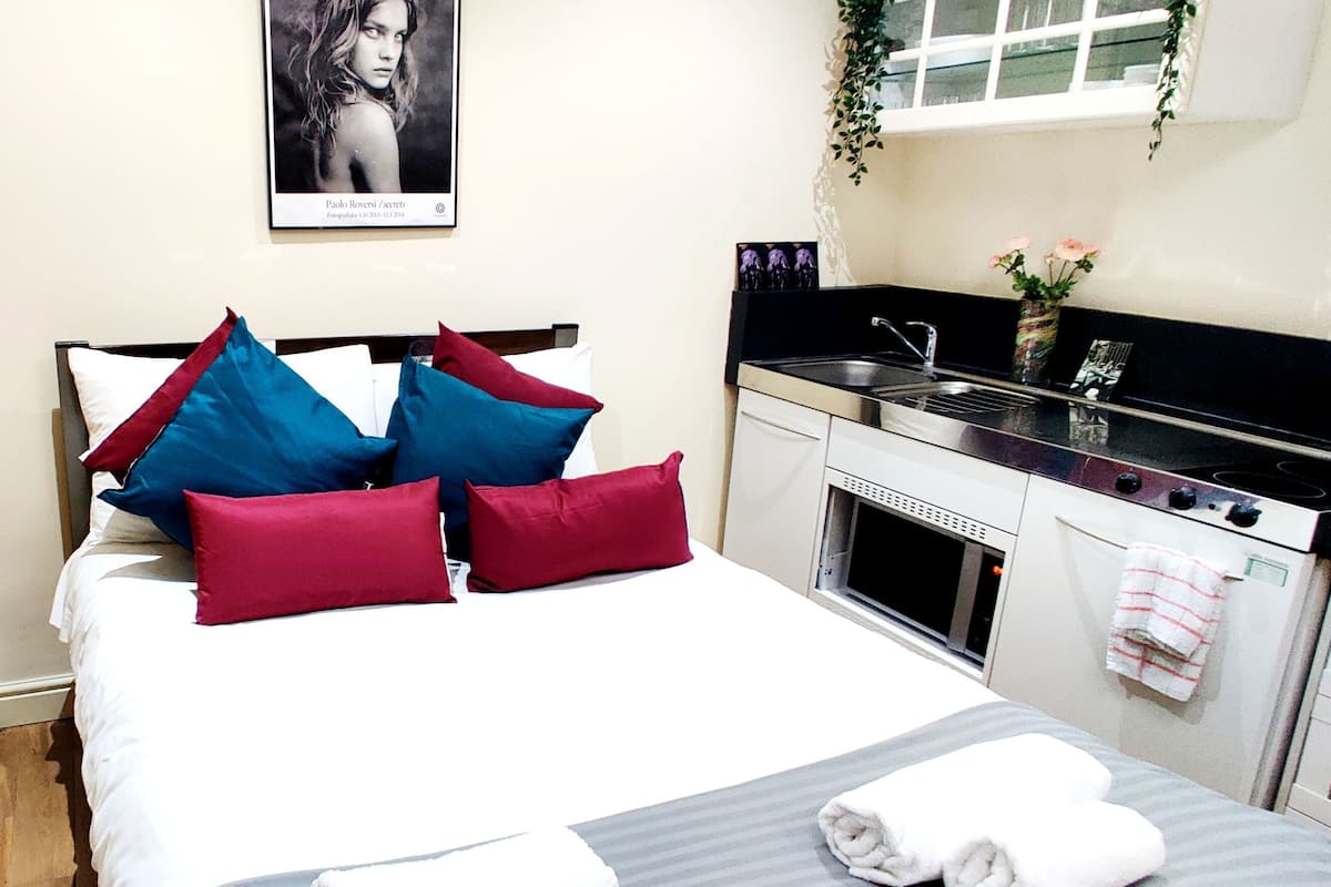 A bedroom with a bed, pillows, a nightstand, a vase with flowers, a framed picture on the wall, a window with a plant, and a kitchenette with a stove and sink.